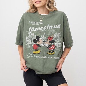 May include: Olive green t-shirt featuring Mickey and Minnie Mouse with the text "Disneyland California Est. 1955" and "The happiest place on earth". The graphic design includes fireworks and a park scene.