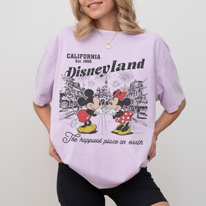 May include: A light purple t-shirt featuring a Disneyland graphic. The graphic includes Mickey and Minnie Mouse, fireworks, and the text "CALIFORNIA Est. 1955 Disneyland The happiest place on earth."