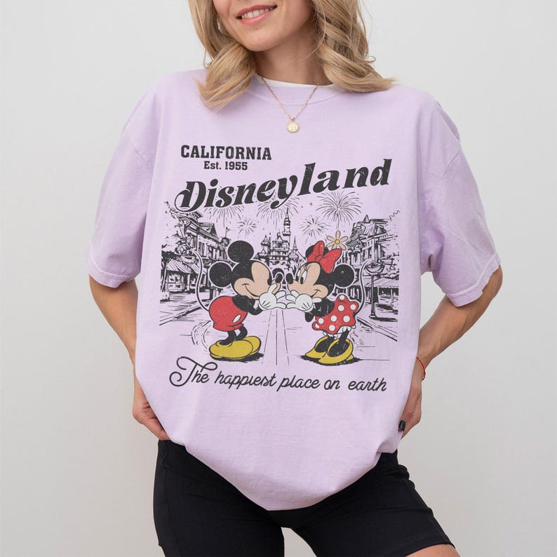 May include: A light purple t-shirt featuring a Disneyland graphic. The graphic includes Mickey and Minnie Mouse, fireworks, and the text "CALIFORNIA Est. 1955 Disneyland The happiest place on earth."