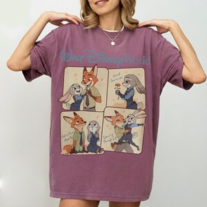 May include: A mauve t-shirt with a graphic of Nick Wilde and Judy Hopps from Zootopia. The shirt has the text "Walt Disney World" above the graphic and the phrases "Sweet Surrender", "Adventure Awaits", and "Dancing Through Life".