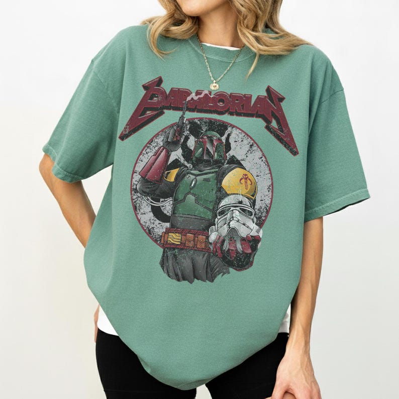 May include: A seafoam green t-shirt featuring a graphic of Boba Fett from Star Wars, with the text "Boba Moria" in a distressed, red font. The design includes a circular background with a vintage aesthetic.