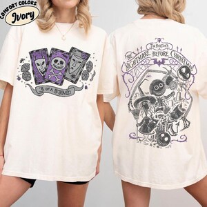 May include: Ivory-colored t-shirt featuring a graphic from "The Nightmare Before Christmas." The front displays three playing cards with character faces and the text "3 of a Kind." The back has a large illustration with the film's title and characters.
