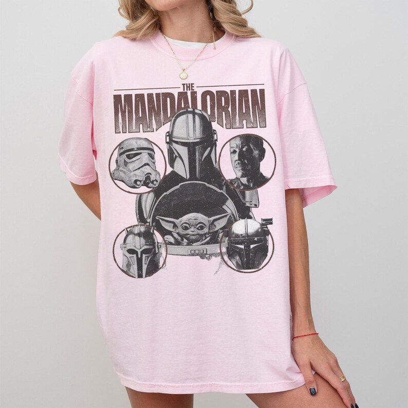 Pu&ograve; includere: Una maglietta rosa chiaro con una grafica di "The Mandalorian". Il design include il titolo dello show e immagini di personaggi come il Mandaloriano, Baby Yoda e uno Stormtrooper. La maglietta ha maniche corte e girocollo.