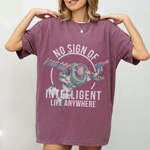 Op de afbeelding: Een bordeauxrood T-shirt met een afbeelding van Buzz Lightyear uit Toy Story. Het ontwerp bevat de tekst "NO SIGN OF INTELLIGENT LIFE ANYWHERE" in het wit. Het shirt heeft korte mouwen en een relaxte pasvorm.