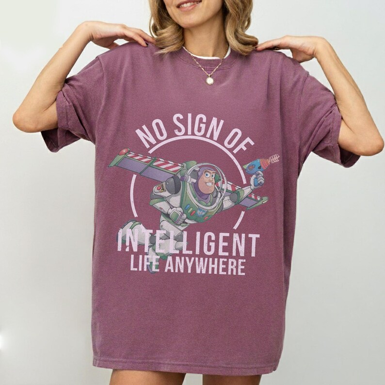 Op de afbeelding: Een bordeauxrood T-shirt met een afbeelding van Buzz Lightyear uit Toy Story. Het ontwerp bevat de tekst "NO SIGN OF INTELLIGENT LIFE ANYWHERE" in het wit. Het shirt heeft korte mouwen en een relaxte pasvorm.