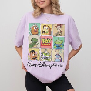 May include: A light purple t-shirt featuring a grid of Toy Story character illustrations. The shirt includes images of Mr. Potato Head, Slinky Dog, Woody, Rex, Buzz Lightyear, and others. The words "Toy Story" and "Walt Disney World" are also printed on the shirt.