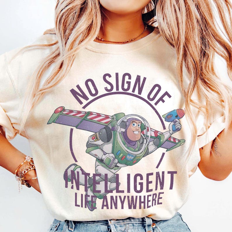 Op de afbeelding: Cr&egrave;mekleurig T-shirt met een afbeelding van Buzz Lightyear uit Toy Story, met de tekst "NO SIGN OF INTELLIGENT LIFE ANYWHERE". Buzz is afgebeeld in zijn ruimtepak, met een straalwapen.
