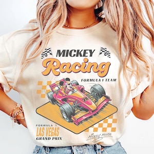 May include: Cream-colored t-shirt featuring a vibrant graphic of Mickey Mouse in a Formula 1 race car. The design includes the text "MICKEY Racing" and "LAS VEGAS GRAND PRIX". The car is pink and yellow, with the number 28.