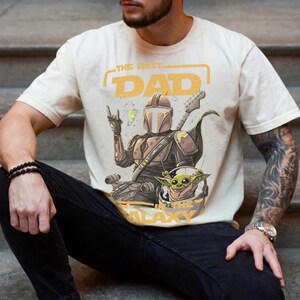 Pu&ograve; includere: T-shirt color crema con una grafica di una figura simile a un Mandaloriano che suona la chitarra, con Grogu in un casco. Il testo recita "THE BEST DAD IN THE GALAXY" in giallo. La maglietta &egrave; indossata da una persona.