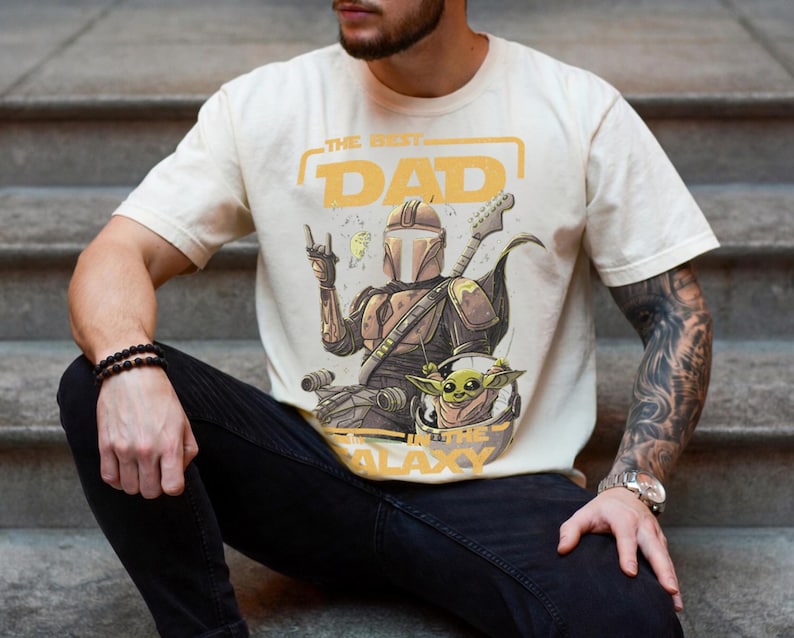 Pu&ograve; includere: T-shirt color crema con una grafica di una figura simile a un Mandaloriano che suona la chitarra, con Grogu in un casco. Il testo recita "THE BEST DAD IN THE GALAXY" in giallo. La maglietta &egrave; indossata da una persona.