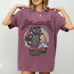 May include: A maroon t-shirt featuring a graphic of Boba Fett from Star Wars, with the word "BAMMORIA" in a distressed font above the image. The design includes a circular background with a vintage aesthetic.