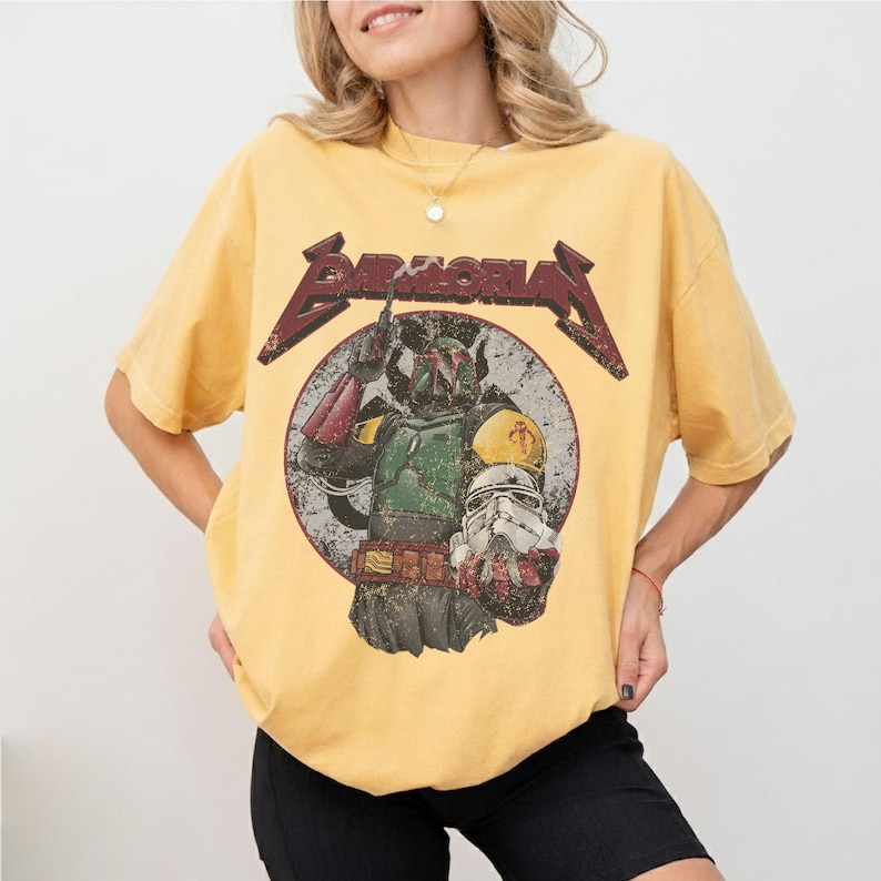 May include: Yellow t-shirt featuring a graphic of Boba Fett and a Stormtrooper helmet inside a circle, with the word "BAMORIAN" in a distressed, red font. The shirt has a vintage, worn look.