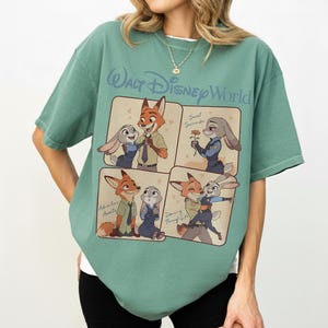May include: A green t-shirt with a Walt Disney World design. The shirt displays four panels with illustrations of Zootopia characters Nick Wilde and Judy Hopps. Text includes "Adventure Awaits", "Sweet Surrender", and "Dancing Through Life".