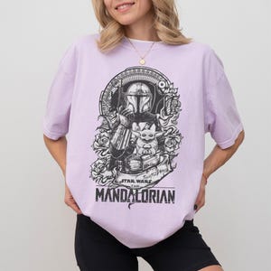 May include: A light purple t-shirt featuring a black and white graphic of The Mandalorian and Grogu from Star Wars. The design includes roses and the text "STAR WARS THE MANDALORIAN".