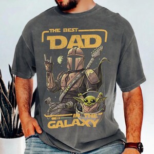 Pu&ograve; includere: T-shirt grigio scuro con una grafica di un personaggio Mandaloriano che suona la chitarra, con Grogu in una capsula fluttuante. Il testo recita "THE BEST DAD IN THE GALAXY" in giallo. Il design include pianeti e stelle.