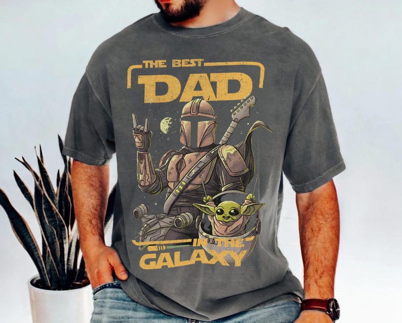 Pu&ograve; includere: T-shirt grigio scuro con una grafica di un personaggio Mandaloriano che suona la chitarra, con Grogu in una capsula fluttuante. Il testo recita "THE BEST DAD IN THE GALAXY" in giallo. Il design include pianeti e stelle.