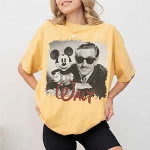K&ouml;nnte beinhalten: Senfgelbes T-Shirt mit einem Schwarz-Wei&szlig;-Bild von Walt Disney und Mickey Mouse. Das Wort "WALT" ist in roter Schrift unter dem Bild geschrieben. Das T-Shirt hat kurze &Auml;rmel und einen Rundhalsausschnitt.