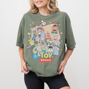 Puede incluir: Una camiseta verde oliva con un gr&aacute;fico colorido de personajes de la pel&iacute;cula animada "Toy Story". El dise&ntilde;o incluye a Woody, Buzz Lightyear y otros personajes queridos. Las palabras "Toy Story" se muestran en una fuente vibrante.