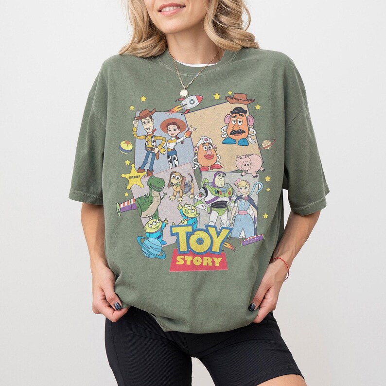 Puede incluir: Una camiseta verde oliva con un gr&aacute;fico colorido de personajes de la pel&iacute;cula animada "Toy Story". El dise&ntilde;o incluye a Woody, Buzz Lightyear y otros personajes queridos. Las palabras "Toy Story" se muestran en una fuente vibrante.