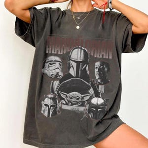 Pu&ograve; includere: T-shirt grigio scuro con una grafica dei personaggi di "The Mandalorian". Il design include il testo "THE MANDALORIAN" sopra le immagini di uno Stormtrooper, del Mandaloriano, di Grogu e di altri personaggi. La maglietta ha un aspetto vintage.