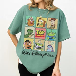 May include: A green t-shirt featuring a grid of Toy Story characters, including Woody, Buzz Lightyear, and Mr. Potato Head. The shirt has the words "Toy Story" and "Walt Disney World" printed on it.