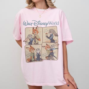 May include: Pale pink t-shirt with a Walt Disney World logo and Zootopia character illustrations. The design includes four panels showing Nick Wilde and Judy Hopps, with text such as "Sweet Surrender" and "Dancing Through Life".