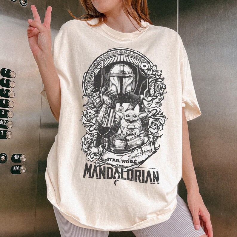 May include: Cream-colored t-shirt featuring a black and white graphic of The Mandalorian and Grogu from Star Wars. The design includes floral elements and the text "STAR WARS THE MANDALORIAN". The shirt has a relaxed fit.