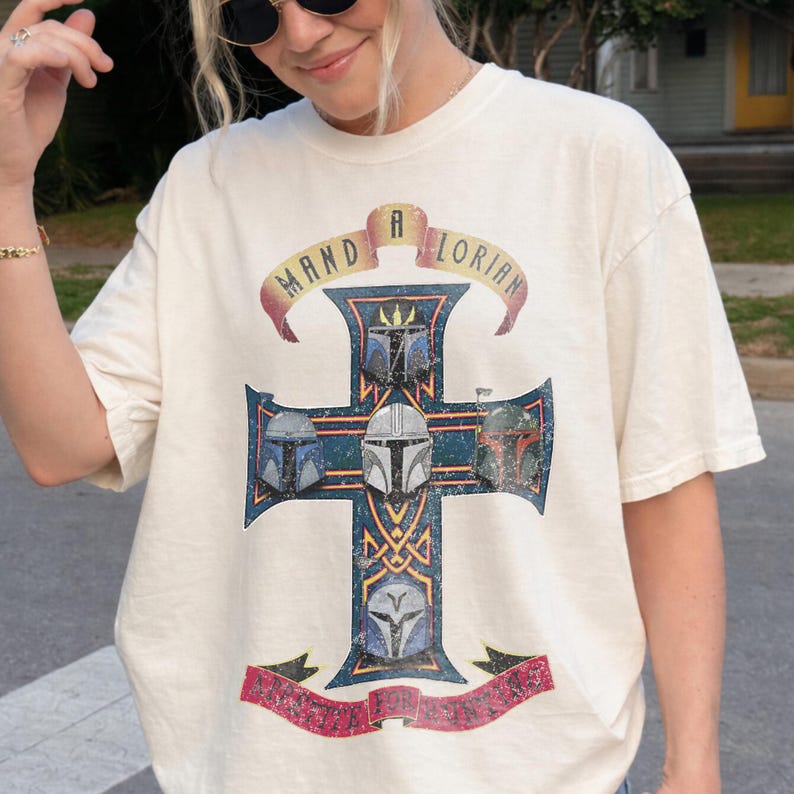 以下が含まれることがあります： オフホワイトの半袖Tシャツ。5つのマンダロリアン・ヘルメットが描かれた大きな青い十字架のグラフィックが特徴です。十字架の上には「MAND A LORIAN」と書かれたバナーがあり、下部にもテキスト入りのバナーが配置されています。全体的にヴィンテージ風の掠れたデザインです。
