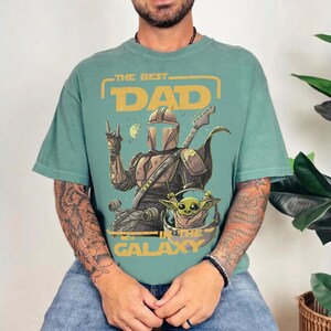 Pu&ograve; includere: Una maglietta color verde acqua con una grafica raffigurante un personaggio in stile Mandaloriano che suona la chitarra, con Grogu. Il testo recita "THE BEST DAD IN THE GALAXY" in giallo e arancione.