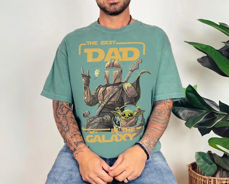 Pu&ograve; includere: Una maglietta color verde acqua con una grafica raffigurante un personaggio in stile Mandaloriano che suona la chitarra, con Grogu. Il testo recita "THE BEST DAD IN THE GALAXY" in giallo e arancione.