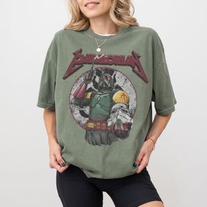 May include: Olive green t-shirt featuring a distressed graphic of a character from a science fiction film. The graphic includes the text "BAMBORIA" in a stylized font. The character is depicted in a circle with a white background.