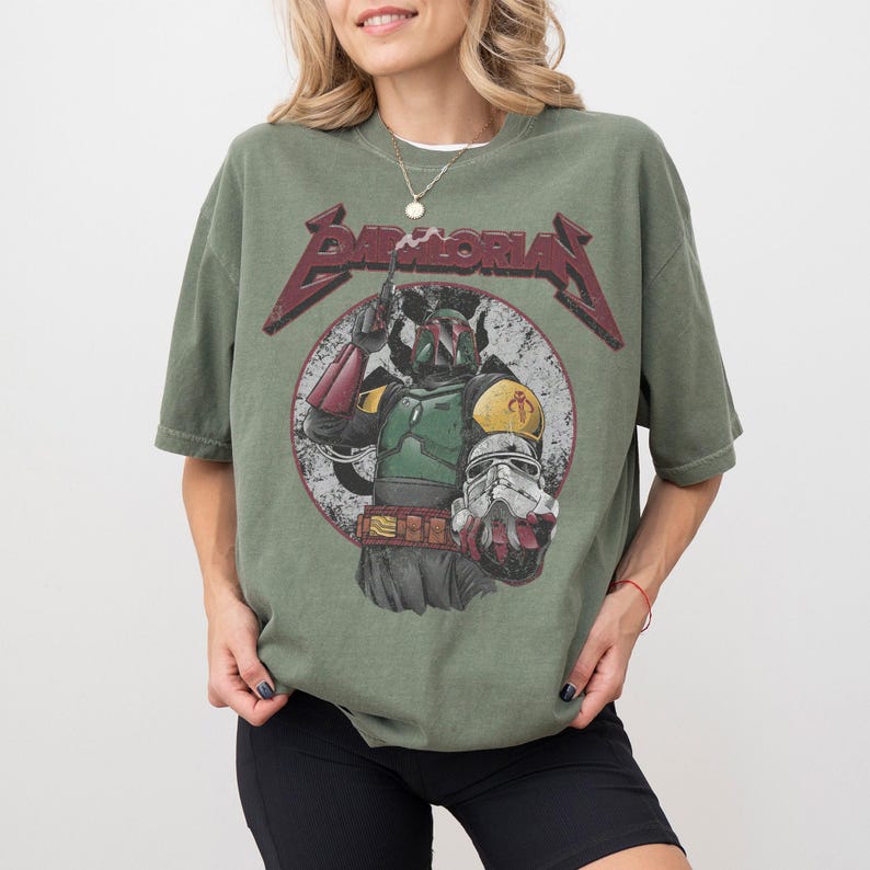 May include: Olive green t-shirt featuring a distressed graphic of a character from a science fiction film. The graphic includes the text "BAMBORIA" in a stylized font. The character is depicted in a circle with a white background.