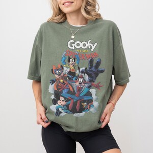 Op de afbeelding: Olijfgroen T-shirt met een cartoonafbeelding van Goofy als Spider-Man en andere personages. De tekst "Goofy became Spider-Man" staat boven de personages gedrukt. Het shirt heeft korte mouwen en een ronde hals.