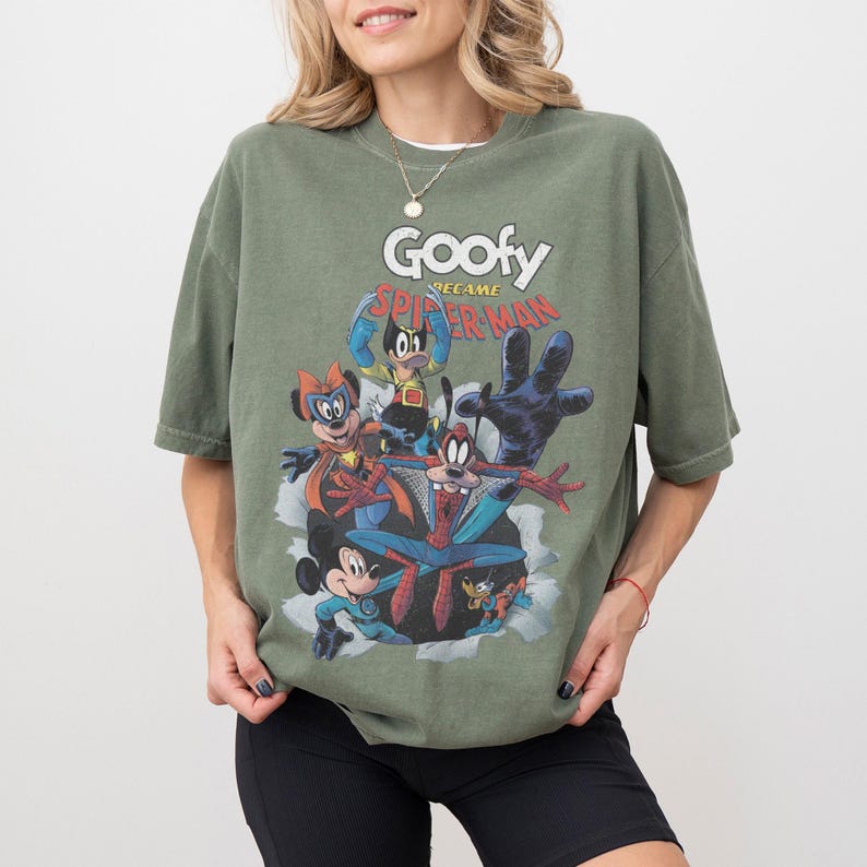 Op de afbeelding: Olijfgroen T-shirt met een cartoonafbeelding van Goofy als Spider-Man en andere personages. De tekst "Goofy became Spider-Man" staat boven de personages gedrukt. Het shirt heeft korte mouwen en een ronde hals.