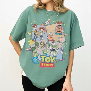 Puede incluir: Camiseta verde con un gr&aacute;fico colorido de personajes de la pel&iacute;cula "Toy Story". El dise&ntilde;o incluye a Woody, Buzz Lightyear y otros personajes populares, con las palabras "TOY STORY" en negrita.