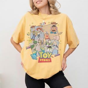 Puede incluir: Camiseta amarilla mostaza con un estampado gr&aacute;fico de personajes de la pel&iacute;cula "Toy Story". El dise&ntilde;o incluye a Woody, Buzz Lightyear y otros personajes, con el t&iacute;tulo de la pel&iacute;cula en rojo y amarillo. La camiseta es de estilo casual, cuello redondo.