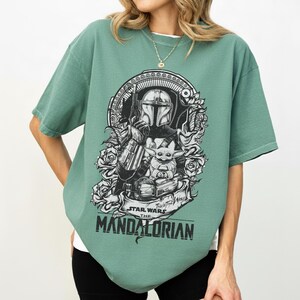 May include: Green short-sleeve t-shirt featuring a black and white graphic of The Mandalorian and Grogu from Star Wars. The design includes floral elements and the text "STAR WARS THE MANDALORIAN".