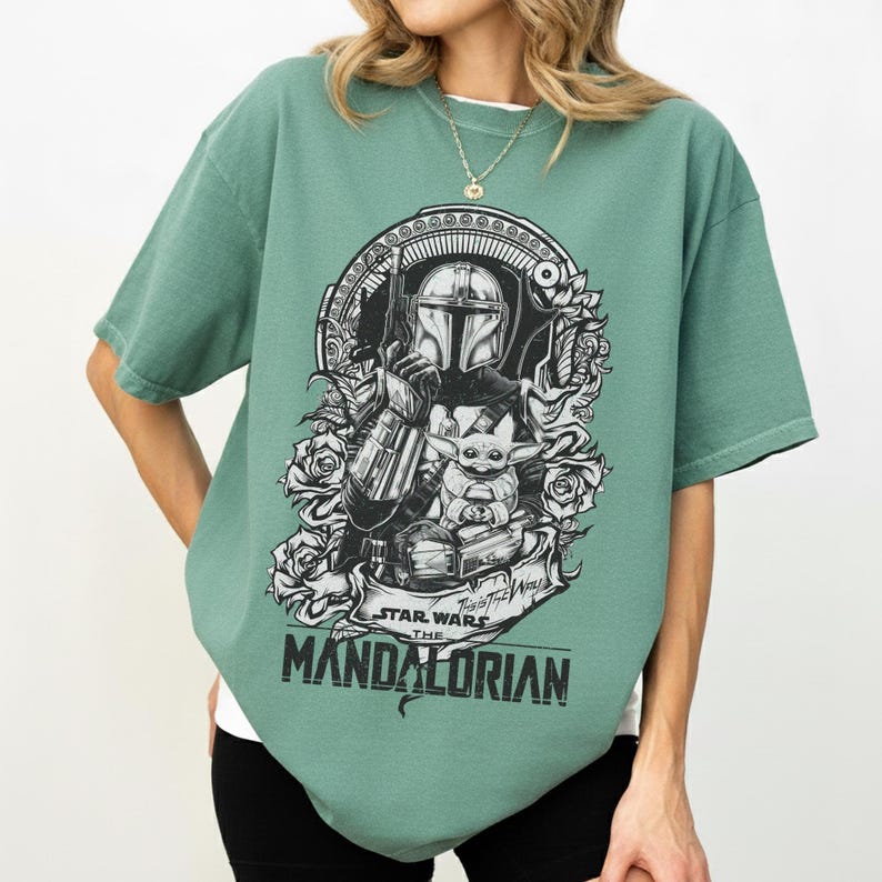 May include: Green short-sleeve t-shirt featuring a black and white graphic of The Mandalorian and Grogu from Star Wars. The design includes floral elements and the text "STAR WARS THE MANDALORIAN".