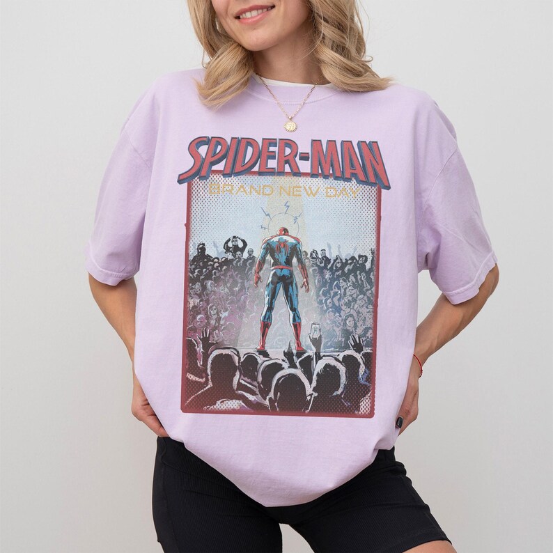 May include: A lilac t-shirt with a Spider-Man graphic. The design shows Spider-Man in front of a crowd, with the text "SPIDER-MAN" and "AND NEW DAY". The graphic has a retro, vintage style.