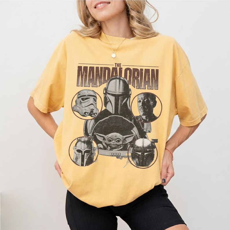Pu&ograve; includere: T-shirt giallo senape con grafica di "The Mandalorian". Il design include il titolo dello show e immagini di personaggi come il Mandaloriano, uno Stormtrooper e Grogu. La maglietta ha un girocollo e maniche corte.