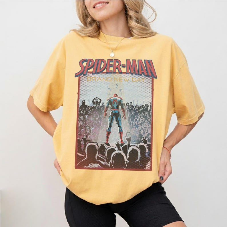 May include: Mustard yellow t-shirt with a Spider-Man graphic. The graphic shows Spider-Man standing before a crowd, with the words "Spider-Man" and "Brand New Day" in a red and white font.