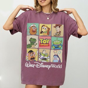 May include: A maroon t-shirt featuring a grid of Toy Story characters, including Mr. Potato Head, Slinky Dog, Woody, Rex, and Buzz Lightyear. The shirt also displays the text "TOY STORY" and "Walt Disney World" in a white font.