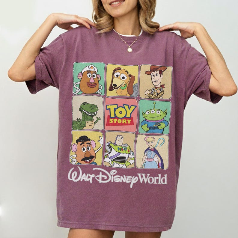 May include: A maroon t-shirt featuring a grid of Toy Story characters, including Mr. Potato Head, Slinky Dog, Woody, Rex, and Buzz Lightyear. The shirt also displays the text "TOY STORY" and "Walt Disney World" in a white font.