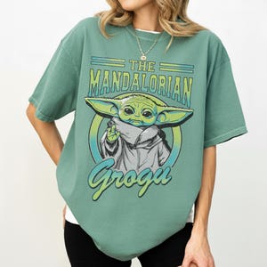 May include: Green t-shirt featuring a graphic of Grogu from The Mandalorian. The design includes the text "The Mandalorian" and "Grogu" in a stylized font. The image of Grogu is in the center of the shirt.