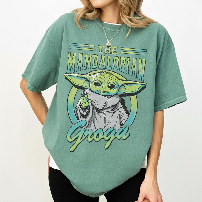 May include: Green t-shirt featuring a graphic of Grogu from The Mandalorian. The design includes the text "The Mandalorian" and "Grogu" in a stylized font. The image of Grogu is in the center of the shirt.