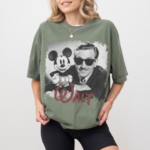 K&ouml;nnte beinhalten: Olivgr&uuml;nes T-Shirt mit einem Schwarz-Wei&szlig;-Bild von Mickey Mouse und Walt Disney. Das Bild hat einen Used-Look mit dem Namen "WALT" in roter Schrift unten. Das T-Shirt hat eine lockere Passform.