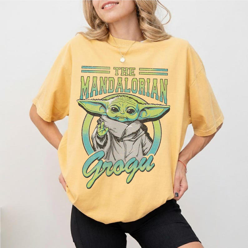 May include: Yellow t-shirt featuring a graphic of Grogu from The Mandalorian. The design includes the text "The Mandalorian" and "Grogu" in green and blue. The shirt is a light yellow color.