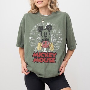 May include: Olive green t-shirt featuring a graphic of Mickey Mouse with a red and yellow color scheme. The words "Mickey Mouse" are printed in red block letters. The shirt has short sleeves and a relaxed fit.