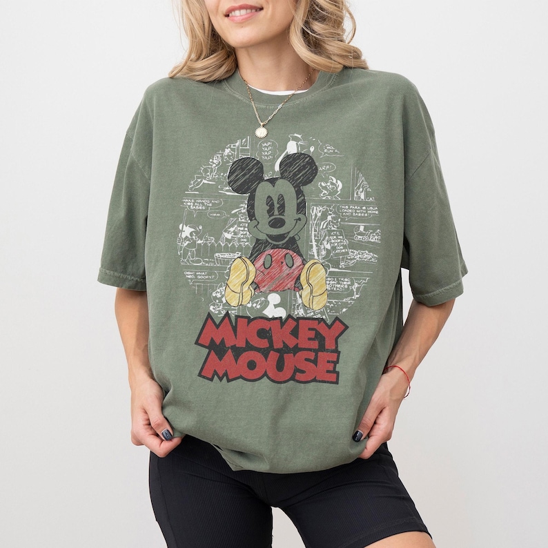 May include: Olive green t-shirt featuring a graphic of Mickey Mouse with a red and yellow color scheme. The words "Mickey Mouse" are printed in red block letters. The shirt has short sleeves and a relaxed fit.