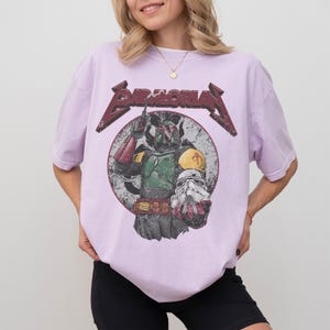 May include: A light purple t-shirt featuring a graphic of a character from the Star Wars franchise. The graphic includes the text "BAMORIAN" in a distressed, metallic style, and a circular image of the character holding a helmet.